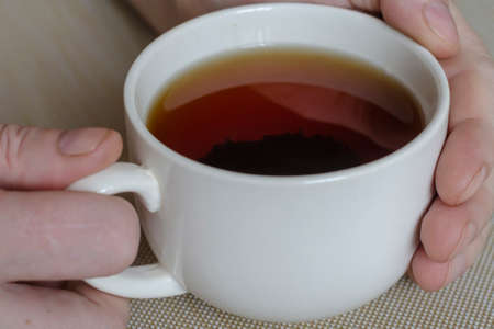 Close-up of male hands and a mug with tea on a beige surface. White ceramic mug full of black tea. The brewed tea leaves lie at the bottom of the mug. Selective focus.の写真素材