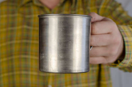 The hand of an adult man in a yellow-green checkered shirt is holding a metal mug. Middle-aged man, Caucasian, indoors. Close-up, selective focus.の写真素材