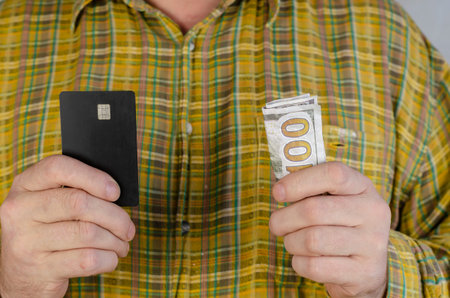 An adult man in a green and yellow plaid shirt holds money and a bank card at chest level. Male...