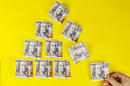 A hand pulls out a dollar banknote from the money pyramid on a yellow background. The crumbling triangular pyramid shape of one hundred American dollars.の写真素材