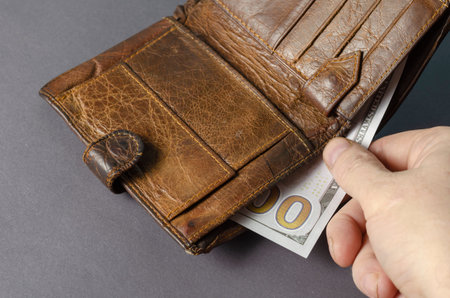A hand pulls out a dollar bill from a brown leather wallet on a gray background. One hundred...