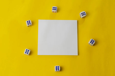 Old dice and white blank paper on a yellow background. Six random white dice and a square sticky note sticker. Top view at an angle. Selective focus.の写真素材