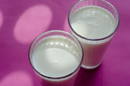 Two full glasses of kefir on a pinky surface with shadows. Transparent glasses with a checkered pattern. Abstract sunny shadows on pink. Selective focus.の写真素材
