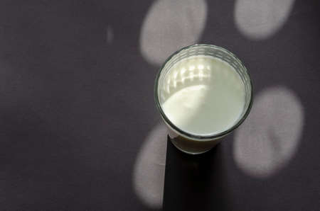 A glass of kefir on a gray surface with shadows. Transparent glass with a checkered pattern. Abstract sunny shadows on gray. Copy space, selective focus.の写真素材