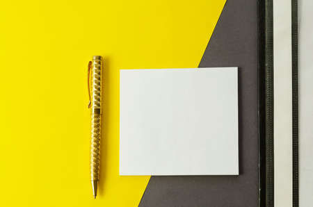 White square blank tear-off paper for notes, binder folder and pen on gray-yellow background. A small stack of paper stickers, a plastic folder and a gold-colored metal pen on a two-tone background. Top view, flat layの写真素材
