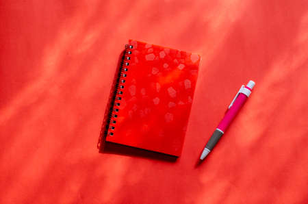 Red closed notepad and pink pen on a red background with shadows. Spring-loaded notebook with plastic cover. Top view at an angle.の写真素材
