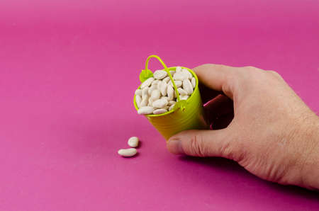 Dry white beans and green bucket and a man's hand on a pink background. A hand empties a legume grain from a small bucket. Close-up, selective focusの写真素材
