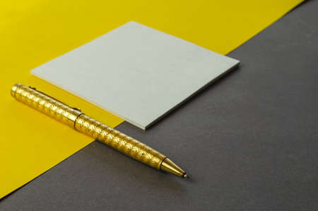 White square blank tear-off note paper and pen on yellow gray background. A small stack of paper stickers and a gold-colored metal pen on a two-tone background. No people.の写真素材