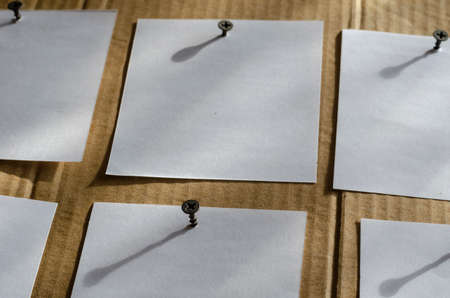 Six Note Paper Stickers are screwed to the brown cardboard with metal screws. White blank square sheets of paper on brown background with shadows.View at an angle. Selective focus.の写真素材