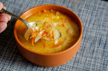A hand with a metal spoon mixes sour cream in a brown earthenware bowl with cabbage soup. A serving...