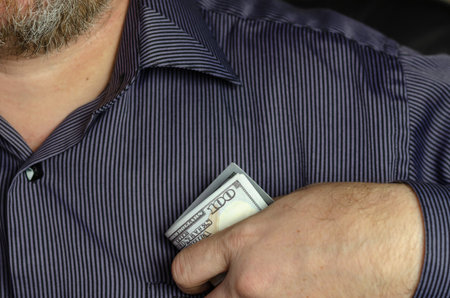 A man in a blue striped shirt puts money in his breast pocket. Hundred dollar bills in the Hand of...