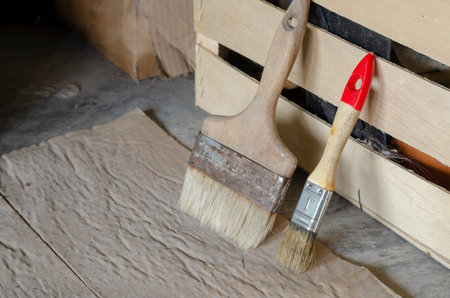 Old paint brushes stand by the wooden box. Two brushes of different sizes stand on the floor covered...