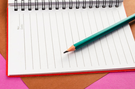 Open spring-loaded notebook and pencil on a pink and brown background. White notepad and green...