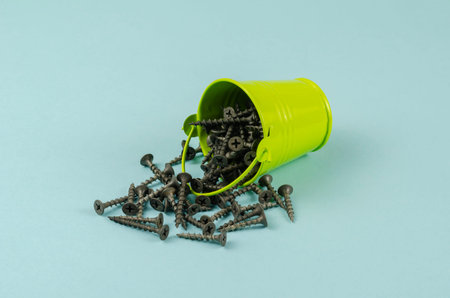 Screws and a small green bucket on a blue background. Black metal hardware spilled out of the fallen...