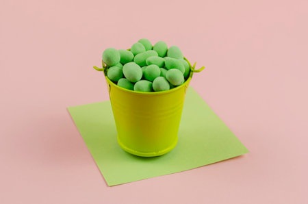 Minimalistic still life with dragees, a bucket and a sheet of paper on a pink background. A metal bucket full of sweets. Square sheet of green paper. Close-up, selective focus.の写真素材