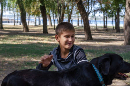 Emotional portrait of a boy stroking a black dog with a blue collar in the park. A 10-year-old child and an 11-year-old female Labrador. A walk in the park with a pet. Blurry hand movement.の写真素材