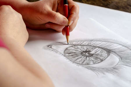 A middle-aged woman draws with a pencil on white paper. An adult woman sits at a wooden table and draws a human eye. Art, hobby, and recreation. Artist's talent. Indoors.の写真素材