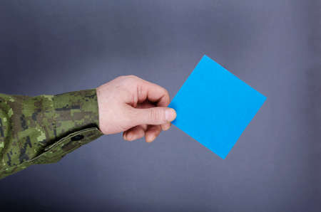 An army officer's hand holds a blue square sheet of paper on a gray background. A middle-aged adult man in camouflage uniform. Military themes, contract military service, concept veterans.の写真素材