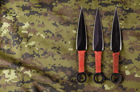 Black Throwing knives on camouflage cloth. Three special knives with red handles crossed. Military theme. View from above at an angle. Copy space.の写真素材