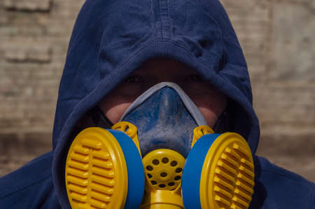 Portrait of an adult unrecognizable man wearing a respirator and hood on against a brick wall. Middle-aged man wearing an industrial blue-and-yellow respirator looks into the camera. Outside. Daytimeの写真素材