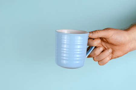 Hand with a mug on a blue background. The man is holding a light blue ceramic mug. Side view.の写真素材
