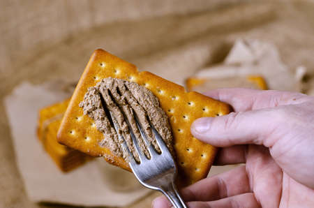 Close-up of a hand spreading pate on a cracker with a fork. A man prepares himself an appetizer with liver pate. High angle view. Selective focus. Homemade food.の写真素材