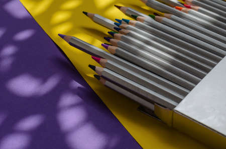 Light and shadow composition with a set of gray wooden pencils on a yellow and blue background. Open-box with multicolored watercolor pencils and paintbrush. View from above at an angle.の写真素材