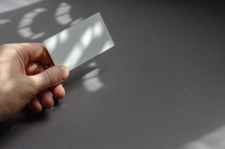 Minimalist light and shade composition of a hand with a business card on a gray background. Man holding an empty paper rectangle in an abstract shadow-light pattern. Advertising, business concept.の写真素材
