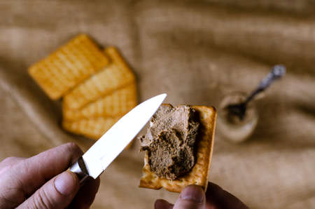Close-up of a hand spreading pate on a cracker with a knife. A man prepares himself an appetizer with liver pate. Crackers and jar in the background. High angle view. Selective focus. Homemade food.の写真素材
