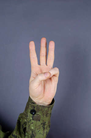 Hand in camouflage shows gesture with his fingers on gray background. Index, middle and ring fingers are raised up. The designation of the number six in the military. Army gesture language.の写真素材