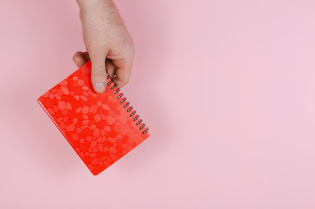 Hand and notepad on pink backdrop. An adult man holds a closed red notebook on a spring. Side view.の写真素材