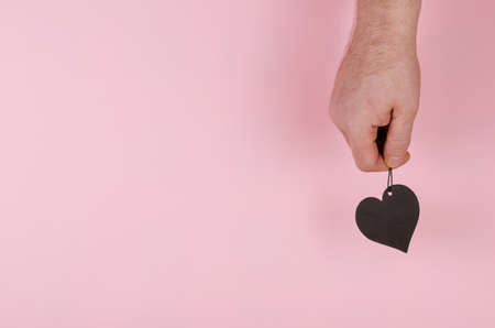 A black heart and a hand on a pink background. Man holding upright a black paper heart. Side view. Copy space.の写真素材