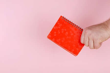 Hand and notepad on pink backdrop. An adult man holds a closed red notebook on a spring. Side view.の写真素材