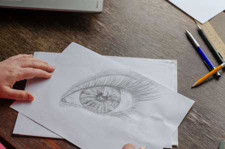 A middle-aged woman draws with a pencil on white paper. An adult woman sits at a wooden table and draws a human eye. Art, hobby, and recreation. Artist's talent. Indoors.の写真素材
