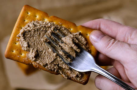 Close-up of a hand spreading pate on a cracker with a fork. A man prepares himself an appetizer with liver pate. High angle view. Selective focus. Homemade food.の写真素材