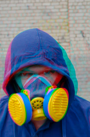 Defocused portrait of an unrecognizable man wearing a hood and a blue and yellow respirator with split glitch effect. An adult man is looking at the camera. Outdoors.の写真素材