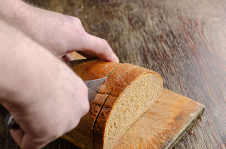 Hands slice bread on a wooden cutting board. A grown man slices a loaf of rye bread with a large knife. Close-up. Selective focus.の写真素材