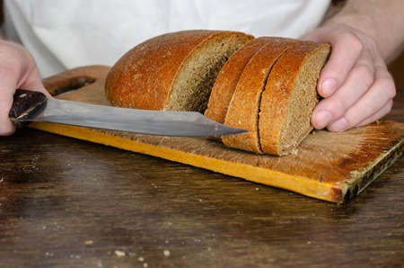 Hands slice bread on a wooden cutting board. A grown man slices a loaf of rye bread with a large knife. Close-up. Selective focus.の写真素材
