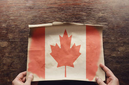 An old, shabby piece of paper with the Canadian flag on it lies on the scratched table. The man's hands hold the unfolded piece of parchment to the brown wooden table. Top viewの写真素材