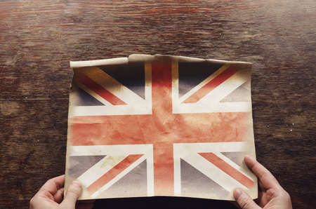 The flag of the United Kingdom of Great Britain and Northern Ireland on a worn piece of paper. A man's hands hold an unfolded piece of parchment on a brown wooden table. Top viewの写真素材