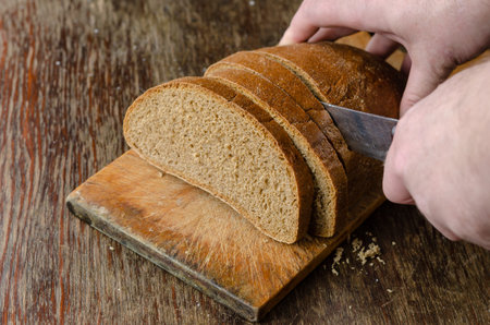 Hands slice bread on a wooden cutting board. A grown man slices a loaf of rye bread with a large...
