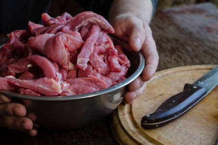 A man's hands hold a full metal bowl of sliced beef. A portion of raw beef stroganoff meat in a metal bowl. Cutting board, knife, mussat.の写真素材