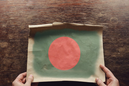 On the scratched table lies an old, tattered piece of paper with a picture of the Bangladeshi flag...