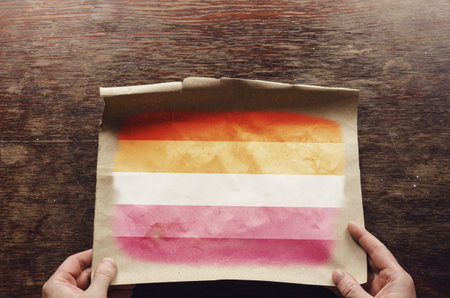 On the scratched table lies an old, tattered piece of paper with a picture of the Flag of lesbian pride. The man's hands press the unfolded piece of parchment against the brown wooden tableの写真素材