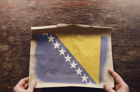 The national flag of Bosnia and Herzegovina on a torn piece of paper. A man's hands press an unfolded sheet of parchment to a brown wooden table. View from aboveの写真素材
