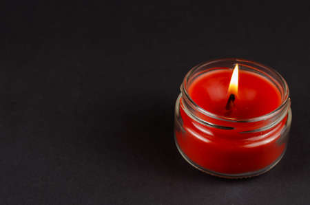 A lit red candle in a glass jar on a black background. The flame of a wax candle in semi-darkness. Side view. Copy space.の写真素材