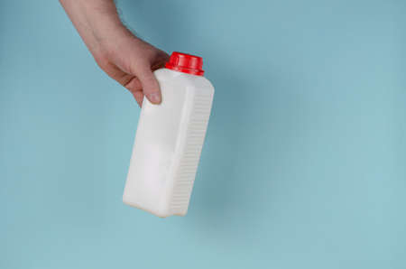 Hand holds white container with lid for liquids on blue background. Rectangular plastic generic bottle with red lid. Side view.の写真素材