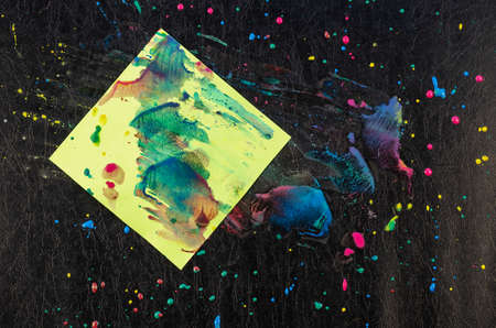 Chaotic drops of paint and piece of paper on black texture. Yellow, red, blue dots and splashes. Green square sheet of paper stained with paints. Abstract multitasking background.の写真素材