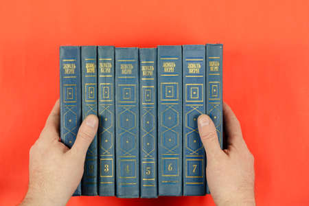 Books of classical literature on a red background. Men's hands hold the Collected Works of Jules Verne in 8 volumes. Hardcover books in Russian. Kiev, Ukraine - 04 27 2021のeditorial素材