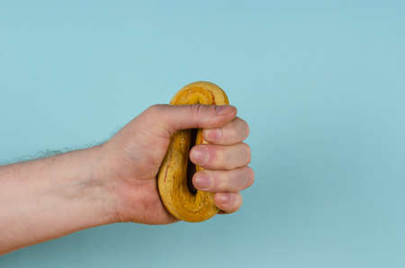The hand is clutching a yellow rubber band on a blue background. Adult male holding a hand-held round hand trainer. Side view. Sports, active lifestyle.の写真素材